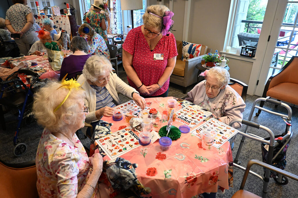Residents gather around for a game of bingo, adorning miniature hats. The room is filled with light, pastel colored decorations.
